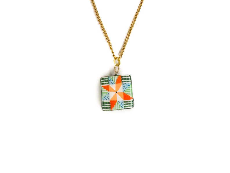 May include: A gold-colored chain necklace with a square pendant. The pendant features a colorful patchwork design with orange, green, and blue geometric shapes. The necklace is displayed against a white background.