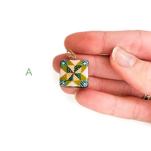 May include: A square pendant with a quilt-inspired design. The pendant features a patchwork pattern in shades of pink, green, blue, and yellow. It has a gold-colored bail for attaching to a chain. The pendant is held in a person's hand.