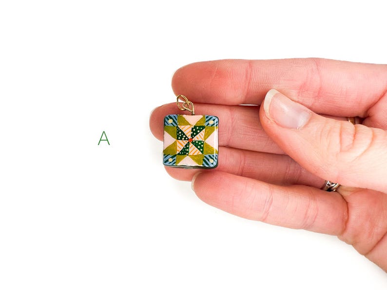 May include: A square pendant with a quilt-inspired design. The pendant features a patchwork pattern in shades of pink, green, blue, and yellow. It has a gold-colored bail for attaching to a chain. The pendant is held in a person's hand.