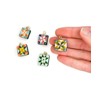 May include: Five square pendants with colorful quilt patterns and gold-colored loops. The pendants feature various geometric designs in shades of green, blue, red, and yellow. The pendants are displayed on a white background.