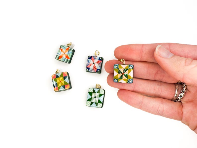 May include: Five square pendants with colorful quilt patterns and gold-colored loops. The pendants feature various geometric designs in shades of green, blue, red, and yellow. The pendants are displayed on a white background.