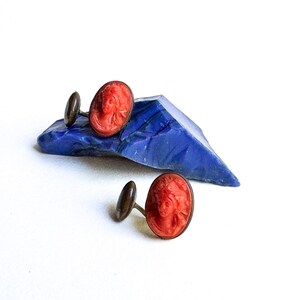 May include: A pair of cufflinks with red cameo insets featuring a woman's face. The cufflinks are displayed on a blue stone.