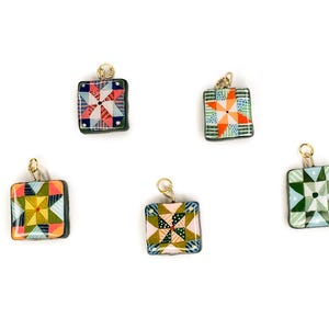 May include: Five square pendants with gold-colored loops. Each pendant features a colorful quilt block design with various geometric patterns in shades of red, blue, green, yellow, and orange. The pendants are set against a white background.