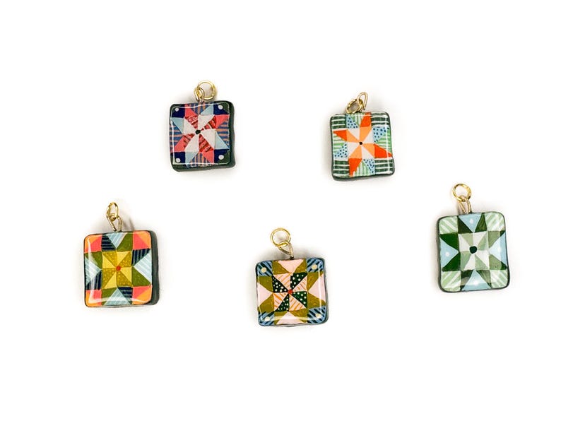 May include: Five square pendants with gold-colored loops. Each pendant features a colorful quilt block design with various geometric patterns in shades of red, blue, green, yellow, and orange. The pendants are set against a white background.