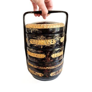 May include: A black and gold tiered lunch box with a handle. The box is decorated with intricate floral patterns and gold accents.