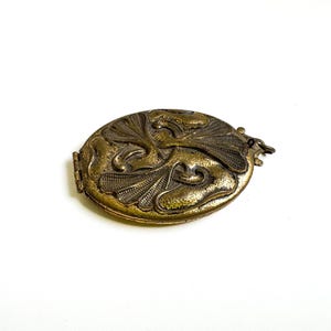 May include: A gold-toned oval locket with an intricate floral design. The locket has a hinged opening and a small loop for attaching a chain.