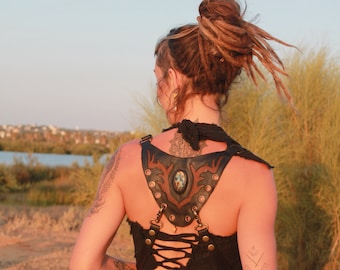 May include: A woman with dreadlocks wears a black top with a leather harness featuring a large gemstone. The harness has a lace-up back and is adorned with metal accents.