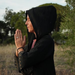 Könnte beinhalten: Eine Person in einer schwarzen Kapuzenjacke mit goldenen Stickereien steht auf einem Feld und hält die Hände zum Gebet gefaltet.