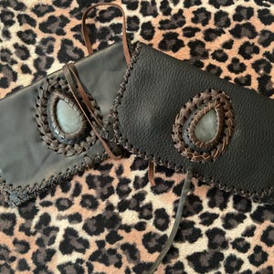 Può includere: Due pochette in pelle nera con una pietra a forma di goccia e finiture in pelle intrecciata. Le borse hanno un bordo cucito e un cinturino in pelle marrone. Lo sfondo è una stampa leopardata.