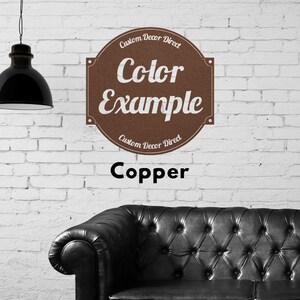 May include: A copper colored sign with the text "Custom Decor Direct" and "Color Example" on a white brick wall. The sign is above a black leather sofa.