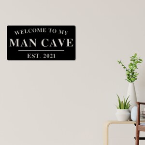 May include: Black metal sign with white text that reads "Welcome to my Man Cave Est. 2021".