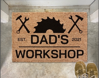 May include: A brown coir doormat with the text "EST. DAD'S 2021 WORKSHOP" in black lettering. The doormat also features a black saw blade and two crossed tools, a hammer and wrench.