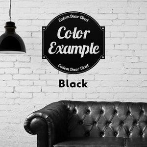 May include: Black and white sign with the text "Custom Decor Direct" and "Color Example" in a circle. The sign is on a white brick wall with a black leather couch in the foreground. The text "Black" is below the sign.