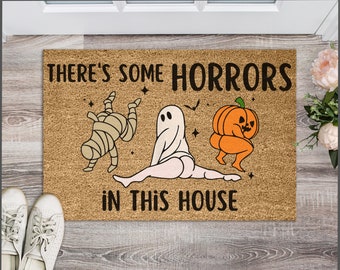 May include: A brown coir doormat with the text "There's some horrors in this house". The text is in black and white. There are three cartoon characters on the mat: a mummy, a ghost, and a pumpkin. The characters are all in a playful, silly pose.