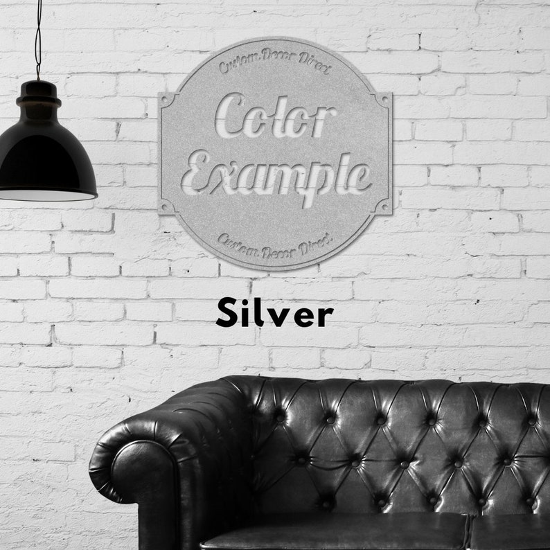 May include: A silver metal sign with the text "Color Example" in a decorative font. The sign is circular and has a raised edge. The sign is hanging on a white brick wall.