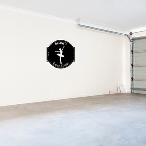 May include: A black and white sign with the text "Kelsey's Dance Studio" and a silhouette of a ballerina in a white dress. The sign is on a white wall in a garage.
