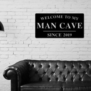 May include: Black metal sign with white text that reads "Welcome to my Man Cave Since 2019". The sign is hanging on a white brick wall above a black leather sofa.