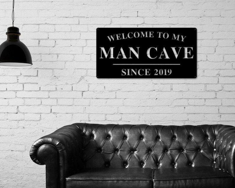 May include: Black metal sign with white text that reads "Welcome to my Man Cave Since 2019". The sign is hanging on a white brick wall above a black leather sofa.