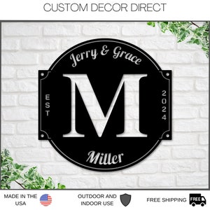 May include: Black metal sign with a white letter M in the center. The sign has a rounded top and a rectangular bottom. The text "Jerry & Grace" is written in a cursive font above the letter M. The text "Miller" is written below the letter M. The text "EST 2024" is written on the right side of the sign.