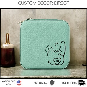 May include: A light green square-shaped jewelry box with a zipper closure. The box is personalized with the name "Nicole" and a stethoscope with a heart that says "RN".