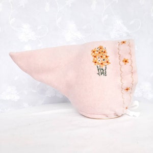Puede incluir: Un gorro de tela rosa con un borde festoneado y flores amarillas bordadas. El gorro está sobre una superficie blanca.
