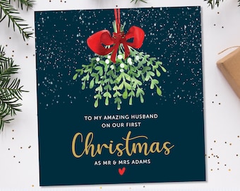May include: A Christmas card with a green mistletoe sprig with a red bow hanging from the top. The card has a dark blue background with white snowflakes and gold text that reads "To my amazing husband on our first Christmas as Mr & Mrs Adams".