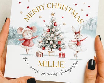 Puede incluir: Tarjeta de Navidad con la inscripción dorada "Merry Christmas". La tarjeta presenta una ilustración en acuarela de un ratón y un conejo, un árbol de Navidad decorado y el nombre "Millie". La tarjeta está sostenida por una persona.