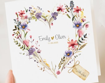 May include: A white card featuring a heart-shaped floral design in pink, purple, and blue. The names "Emily" and "Oliver" are written inside the heart, with the date 11.06.2025. A small tag tied with string reads "With Love."