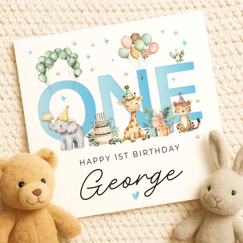 May include: A birthday card with the word "ONE" in light blue, decorated with cartoon animals, balloons, and a cake. The card reads "HAPPY 1ST BIRTHDAY George" in script. A teddy bear and a stuffed bunny are in the foreground.