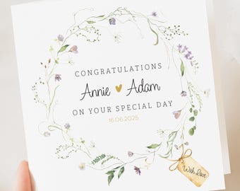 May include: A white wedding congratulations card featuring a floral wreath. The card reads "Congratulations Annie ♥ Adam On Your Special Day 16.06.2025." A small tag with "With Love" is attached. The wreath includes green leaves and purple flowers.