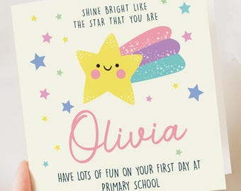 May include: A yellow star with a smiling face and a rainbow tail, surrounded by small stars. The text "Shine bright like the star that you are" is above the star. The name "Olivia" is written in pink below the star. The text "Have lots of fun on your first day at primary school" is below the name.
