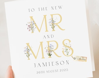 May include: A white wedding card with gold lettering that reads "TO THE NEW MR AND MRS JAMIESON 26TH AUGUST 2025". The letters MR and MRS are decorated with floral accents. A small tag reads "With Love".
