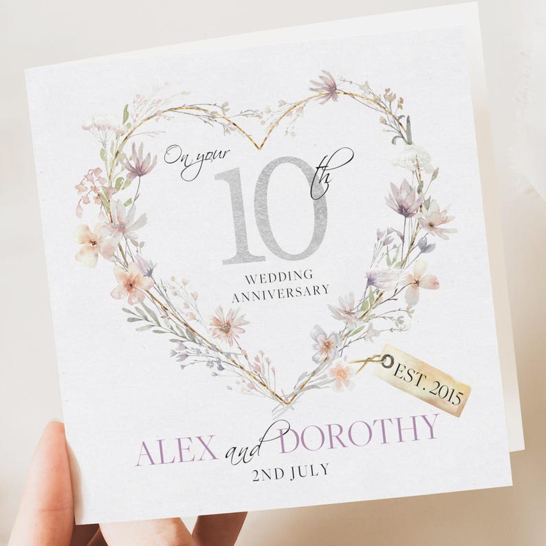 May include: A white card with a floral heart design featuring the number 10 in silver with the text "On your 10th Wedding Anniversary" and a tag that says "Est. 2015". The names "Alex and Dorothy" and the date "2nd July" are printed below the heart.
