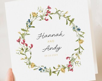 May include: A square wedding invitation with a watercolour floral wreath. The wreath is composed of pink, yellow, and blue flowers with green foliage. The names "Hannah and Andy" are in script, with the date "09.10.2024" below.