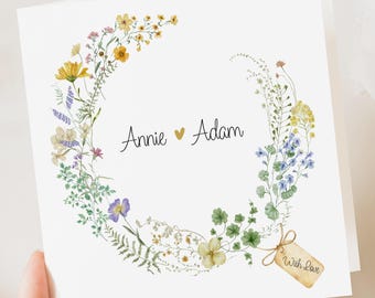 May include: A white greeting card with a watercolour floral wreath in shades of yellow, blue, and purple. The names "Annie" and "Adam" are centered, with a heart symbol between them. A small tag reads "With Love".