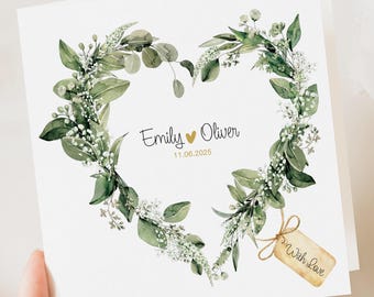Peut inclure: Une carte carrée blanche avec une couronne en forme de cœur de feuilles vertes et de fleurs blanches. Les noms "Emily" et "Oliver" sont écrits en script, avec la date "11.06.2025" en dessous. Une petite étiquette dit "With Love".
