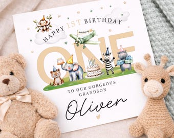 May include: A birthday card with the text "Happy 1st Birthday" and "One" in gold. The card features illustrations of animals, a plane, and clouds. A teddy bear and a giraffe plush toy are next to the card. The card reads "To our gorgeous grandson Oliver."
