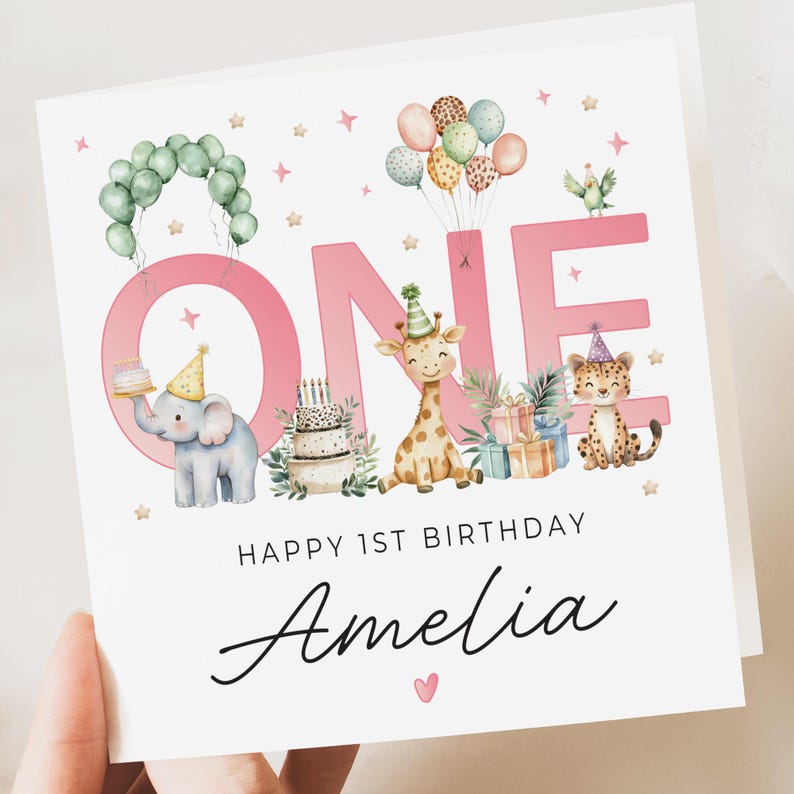 May include: A white greeting card with a pink "ONE" design, featuring cartoon animals, balloons, and a cake. The text "HAPPY 1ST BIRTHDAY" and the name "Amelia" are also present. The card is held by a hand.