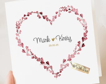 May include: A white card featuring a heart-shaped floral wreath in pink and burgundy. The names "Mark" and "Kirsty" are written in cursive, with the date "29.05.25" below. A small tag reads "With Love".