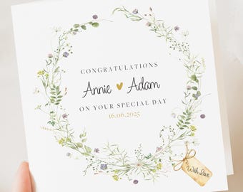 May include: A white wedding card featuring a floral wreath design. The card reads "Congratulations Annie & Adam on your special day 16.06.2025." A small tag tied to the card says "With Love."