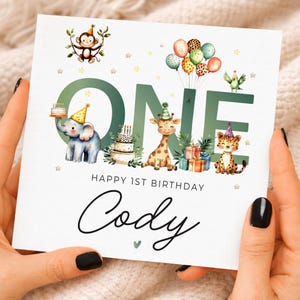 Puede incluir: Una tarjeta de cumpleaños cuadrada con la palabra ONE en letras grandes verdes, decorada con ilustraciones en acuarela de un mono, elefante, jirafa, leopardo y loro con gorros de fiesta, globos, pasteles y regalos, con el texto HAPPY 1ST BIRTHDAY Cody.