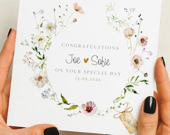 May include: A white wedding congratulations card featuring a floral wreath. The card is inscribed with "Congratulations Joe & Sofie on your special day 13.09.2025." A small tag with "With Love" is attached.