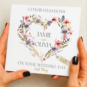 Op de afbeelding: Een witte huwelijkskaart met een bloemenhartontwerp. De kaart toont de namen "Jamie en Olivia" in het hart, met de datum "2nd May" en het jaar "2026". De tekst "Congratulations" staat bovenaan.