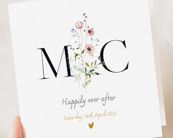 May include: A white wedding card featuring the black initials "M C" and a floral design in pinks, blues, and greens. The words "Happily ever after" and the date "Saturday 16th April 2025" are in gold, with a small heart.