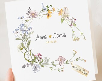 May include: A white wedding card with a watercolour floral wreath of pink, yellow, and blue flowers. The names "Anna" and "James" are in script, with the date "29.05.25". A small tag reads "With Love".