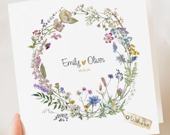 May include: A white card with a floral wreath design. The names "Emily" and "Oliver" are centered within the wreath, with a date below. A small tag reads "With Love".