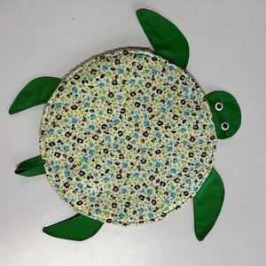 May include: A turtle-shaped fabric item featuring a circular shell with a light green background covered in a small floral pattern of blue and dark brown blossoms. The head, four flippers, and tail are solid green fabric with two googly eyes.