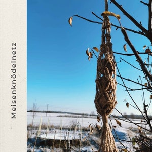 May include: A brown, woven bird feeder hanging from a bare tree branch against a blue sky. The feeder is made of natural twine and has a knotted design. The background shows a snowy field.