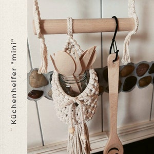 May include: A white macrame hanging organizer with a wooden dowel and a wooden spoon with a smiley face. The organizer is filled with wooden utensils and a glass jar. The text "K&uuml;chenhelfer "mini"" is visible in the image.