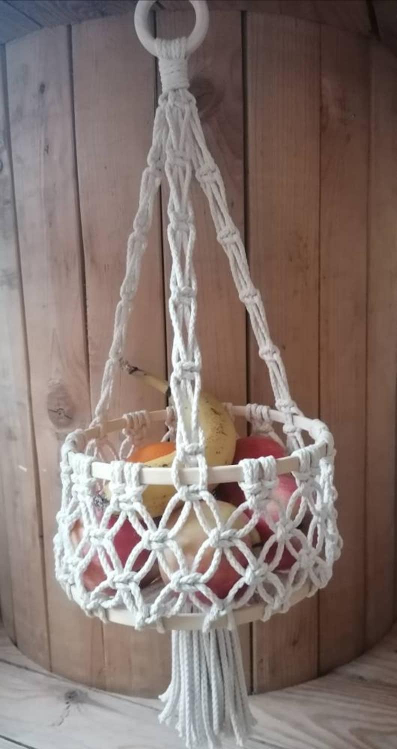 May include: A white macrame hanging basket with a wooden ring at the top and a long tassel at the bottom. The basket is filled with fruit, including apples, pears, and bananas.